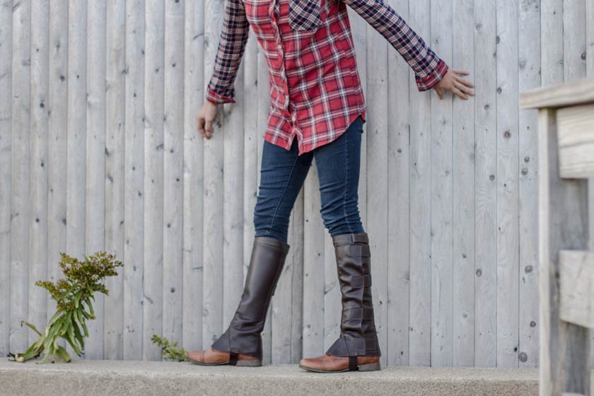 model standing next to wooden fence wearing barnstable ridings brown top grain leather half chaps with velcro straps.