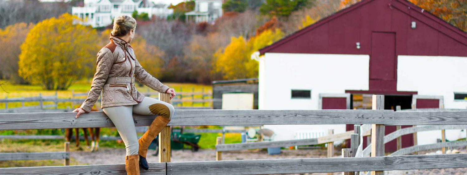 Girl wearing barnstable ridings toast suede half chaps sitting on wooden fence, red and white barn and horse in background. Beautiful fall day and colors on trees and leaves.